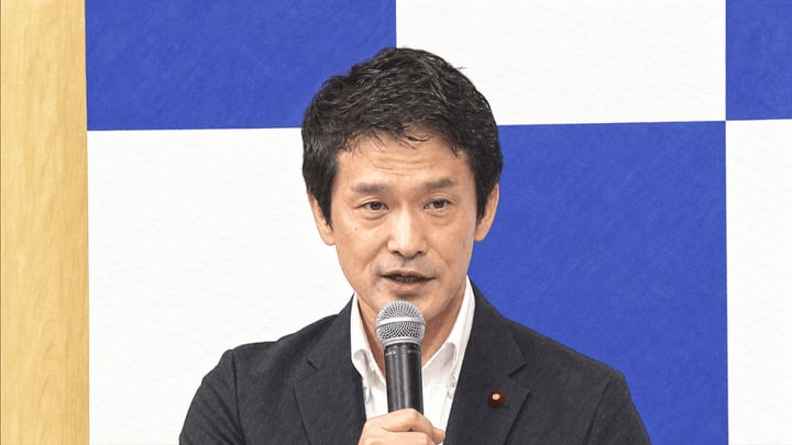 파란색과 흰색 벽 앞에서 검은 재킷을 입은 오가와 준야가 마이크를 손에 들고 발언하는 모습